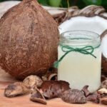 Coconut and coconut oil for natural skincare and nail care, with a glass jar of coconut oil and dried coconut shells on wooden surface, promoting organic beauty treatments.